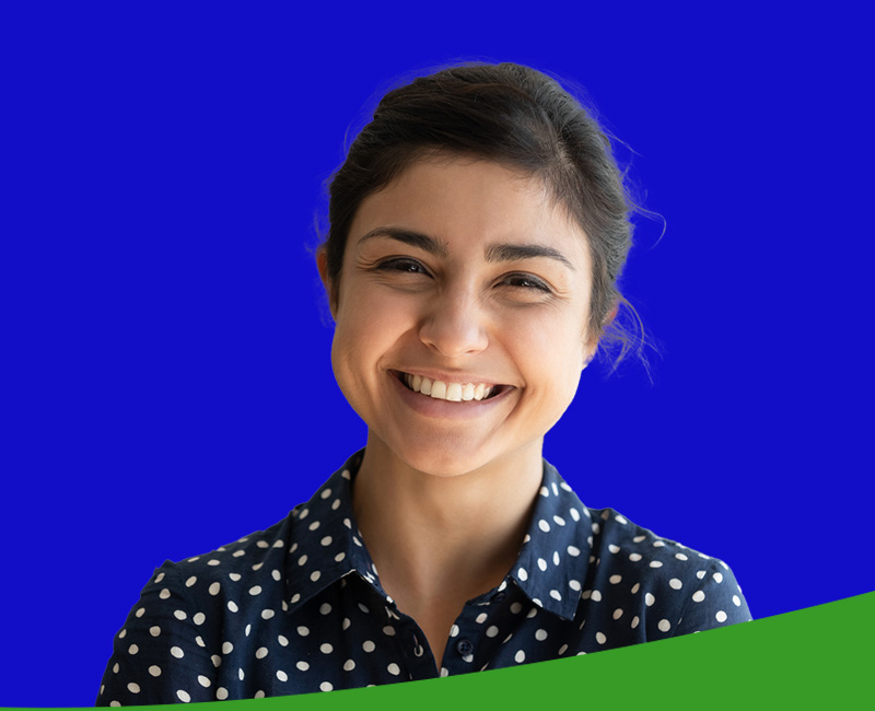 The image shows a person smiling at the camera, wearing a polka dot shirt and a dark blazer, with short hair and standing against a blue background.
