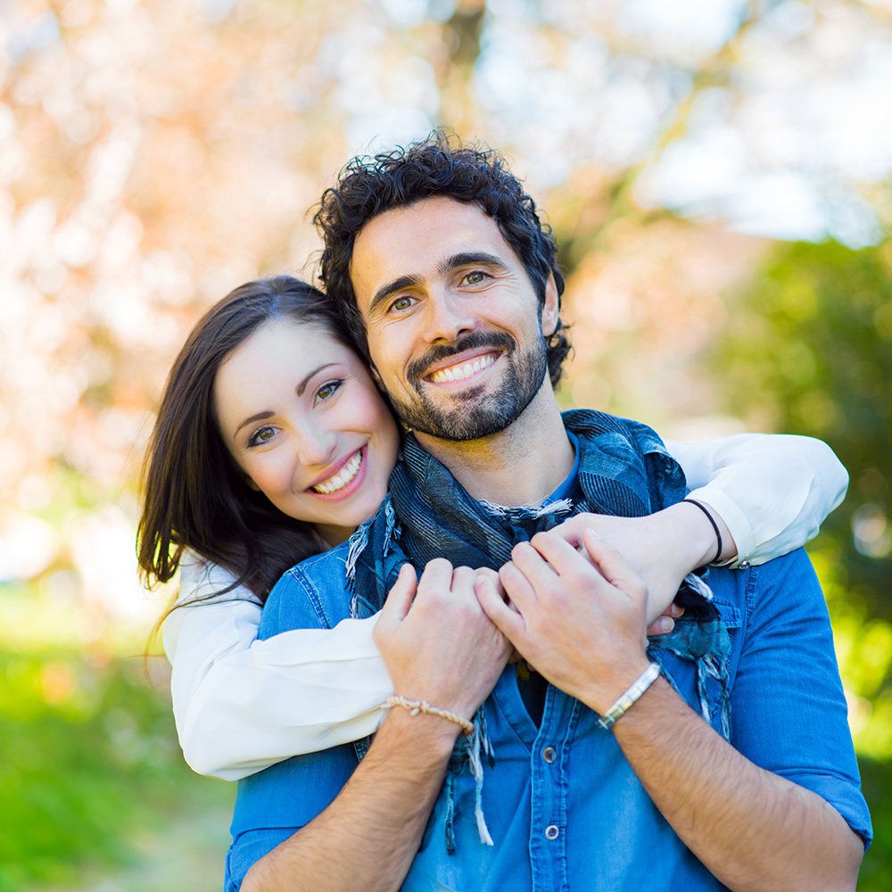 The image shows a man and woman embracing each other outdoors with trees in the background, both smiling at the camera.